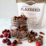 a glass mason jar filled with cinnamon sugar coated nuts, pictured on a white backdrop with red cranberries on the table and a bag of flaxseed behind