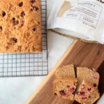 two slices of cran orange bread on a wooden tray with the rest of the loaf on a cooling rack behind